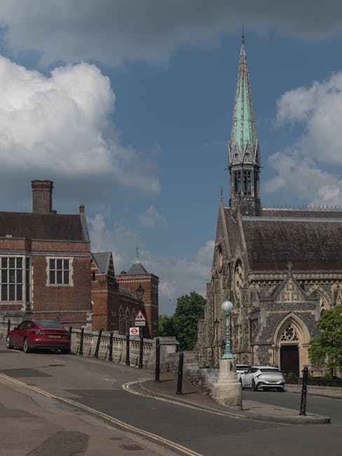 A view of a church with a tall, pointed spire and Gothic architectural details, situated on a street with a gentle incline. In the foreground, there are parked cars, including a red vehicle on the left side and a silver one near the church entrance. The street is lined with black metal bollards and a stone lamppost with a spherical light fixture, which are positioned along the sidewalk and near the church steps. The surrounding buildings are made of brick, with some showing traditional window designs. The sky is partly cloudy with patches of blue visible, indicating daytime. This scene is typical of a historic town area and highlights features relevant to home relocation services such as loading, unloading, and transport within a residential environment, as provided by Man with Van Harrow Weald, specialists in removals.