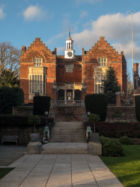 A large historic brick building with a prominent central clock tower topped by a white spire, situated behind a well-maintained garden and stone steps leading up to the entrance. The building features decorative brickwork with stepped gables, tall rectangular windows, and a wooden door at the top of the steps. The surrounding landscape includes trimmed bushes, small trees, and paved walkways, with a clear sky overhead. This setting exemplifies a traditional and heritage-style property suitable for residential home relocation or furniture transport logistics, as handled by Man with Van Harrow Weald, a professional removals service operating in the Harrow Weald area.