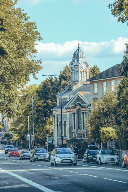 A view of a church with a tall, pointed spire and Gothic architectural details, situated on a street with a gentle incline. In the foreground, there are parked cars, including a red vehicle on the left side and a silver one near the church entrance. The street is lined with black metal bollards and a stone lamppost with a spherical light fixture, which are positioned along the sidewalk and near the church steps. The surrounding buildings are made of brick, with some showing traditional window designs. The sky is partly cloudy with patches of blue visible, indicating daytime. This scene is typical of a historic town area and highlights features relevant to home relocation services such as loading, unloading, and transport within a residential environment, as provided by Man with Van Harrow Weald, specialists in removals.