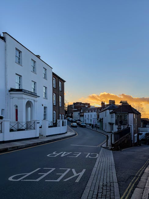 A quiet residential street in Harrow Weald at sunset, featuring a large white multi-storey building with multiple windows, a curved road with a 'KEEP CLEAR' road marking, and a pavement with tactile paving. The road gently slopes uphill and is bordered by traditional terraced houses and low-rise buildings with dark roofs. In the background, there are scattered clouds illuminated by the setting sun, casting a warm glow over the scene. The street appears calm and empty, with no visible vehicles or pedestrians at the moment. This setting provides an ideal environment for house removals and furniture transport services, aligning with the expertise of Man with Van Harrow Weald in relocation logistics and packing and moving activities, captured in a natural and accurate manner.