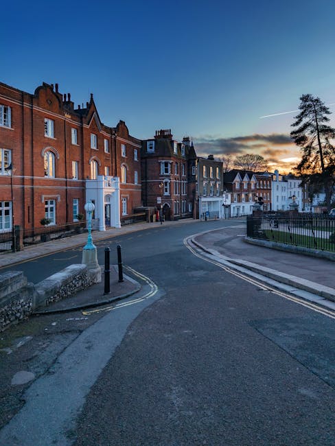 A quiet residential street in the evening with a row of multi-storey brick and white-painted houses on the left side, each featuring bay windows and pitched roofs. The street curves gently to the right, bordered by a low stone wall and lamp posts, with a few pedestrians visible in the distance. On the right, there is a small park area with a black metal railing and a tall, thin tree. The sky shows a sunset with a gradient of orange and blue hues, casting soft light over the scene, suggesting a calm environment suitable for house removals or moving logistics with companies like Man with Van Harrow Weald involved in furniture transport and packing preparations.
