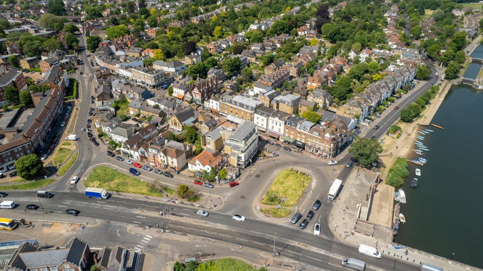 An aerial view of a coastal town in Harrow Weald showing a mixture of multi-storey brick buildings, residential houses with tiled roofs, and greenery. The streets are busy with various vehicles, including cars, vans, and a blue truck, traveling along the main road and around small roundabouts. A section of the waterfront area features a paved promenade with parked boats moored along the water's edge. Inside the town, there are visible parking areas filled with cars and a few pedestrians walking near the buildings. The image captures the urban landscape during daylight with natural lighting, highlighting the typical setting for home relocation activities managed by Man with Van Harrow Weald, where furniture transport and packing and moving processes are integral to local removals services.