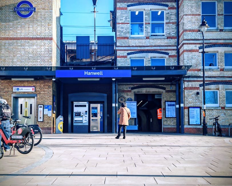 A person dressed in a light brown coat, dark trousers, and carrying a bag, standing outside the entrance of Hanwell station in Harrow Weald, with a blue sign displaying the station name. In front of the station, there are bicycles parked on the pavement, including one with a logo for a bike-sharing service. To the left of the entrance, there are two public ticket machines and an information display panel. The station building is constructed with exposed brickwork and features large windows, with a small balcony or terrace area visible above the entrance. The scene is set during daylight, with bright natural lighting and minimal shadows, representing surroundings where furniture transport and home relocation services by Man with Van Harrow Weald could be coordinated for a smooth moving process.