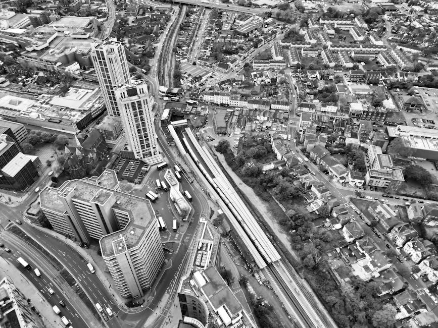 An aerial black-and-white photograph of a dense urban area showcasing a cluster of high-rise buildings, including a prominent tall tower with a modern design and glass windows. The scene captures a busy cityscape with numerous smaller buildings, streets filled with cars, and a network of roads and railway tracks. In the foreground, there are several multi-storey office blocks with flat roofs, some featuring rooftop equipment. The railway lines run diagonally through the image, with platforms visible along the tracks. To the right, a residential neighborhood with houses, trees, and narrow streets is visible. The overall view emphasizes the high-density development typical of a city center, with construction sites, parking areas, and a variety of building types. This image aligns with the context of urban relocations and the logistics of moving within a metropolitan environment, as seen in professional house removals and furniture transport services, such as those offered by Man with Van Harrow Weald.