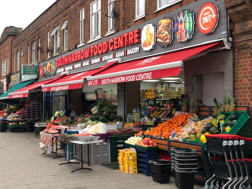The image shows the storefront of South Harrow Food Centre, a local grocery shop with a red and white awning displaying the shop's name and various food categories such as Asian, bakery, and Turkish. In front of the shop, there is a sidewalk market setup with an assortment of fresh produce arranged on tables and in crates, including oranges, watermelons, apples, and leafy greens. Several cardboard boxes and plastic crates are stacked nearby, some used for transporting vegetables and fruits. A small folding table with a floral cloth holds additional items. To the left, there are more display tables with fruit and vegetables, with other shoppers' shopping carts positioned at the edge of the market area. The shop's interior, visible through open shutter doors, shows shelves stocked with packaged goods, indicating a busy local food shop. The building is made of brick, and the scene is lit with natural daylight, highlighting the vibrant colors of the produce and signage. The setup suggests a typical outdoor market scene outside a grocery store, supporting home relocation or moving logistics like packing or transporting household groceries. South Harrow Food Centre is associated with Man with Van Harrow Weald, a local removals service.