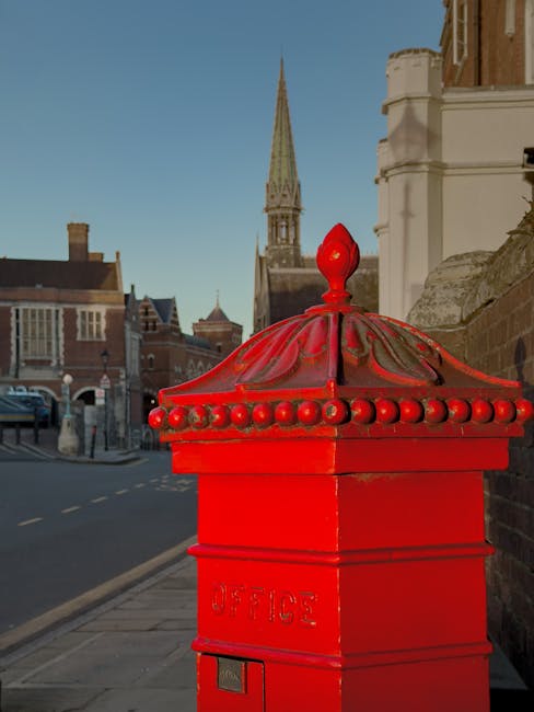 A bright red, traditional British post box with a decorative, domed top and ornamental beads along the edge, is positioned on a pavement next to a stone wall in Harrow Weald. In the background, the scene features a clear blue sky, historic brick buildings, and a prominent church spire, indicating a typical town centre setting. Nearby, there is a parked blue van, potentially used for home relocation or furniture transport services offered by Man with Van Harrow Weald, as part of their house removals and packing and moving operations. The image captures an urban street environment during daylight, highlighting the process of loading, moving, or preparing furniture or boxes for transport, with the post box standing as an iconic local fixture.