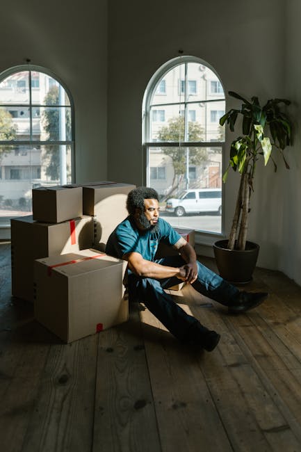 A man with curly black hair and medium skin tone is sitting on a wooden floor inside a room with large, arched windows, which allow natural daylight to illuminate the space. He is dressed in a blue t-shirt and dark jeans, with his arms resting on his knees, appearing to be resting during a home relocation process. Multiple cardboard moving boxes, some sealed with red tape, are stacked nearby, indicating packing activities typical of furniture transport and packing. Behind him, a tall potted plant with broad green leaves is positioned near the window, adding a touch of interior decor. The room’s plain white walls and the natural light coming from outside reveal an urban residential environment with parked cars visible through the windows. This scene captures a moment in the loading or unpacking process during a house removal, with a focus on the interior setting during professional moving services provided by Man with Van Harrow Weald, specializing in home removals and furniture transport.