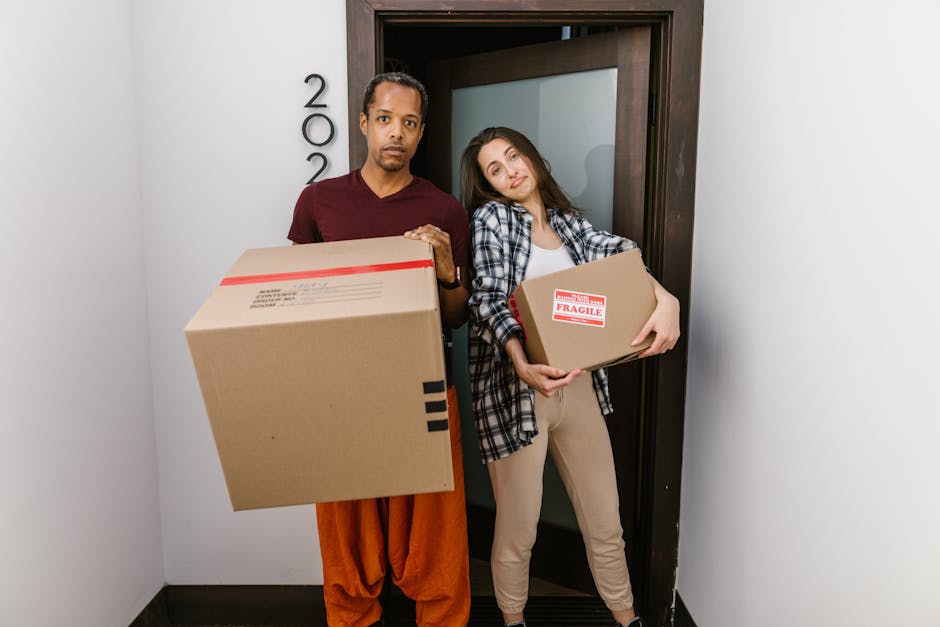 A man and a woman stand inside a doorway, each holding stacked cardboard boxes packed for a home relocation. The man, wearing a maroon shirt and orange trousers, holds a larger box with shipping labels visible on one side, while the woman, dressed in a checkered shirt and beige pants, carries a smaller box marked with a 'Fragile' sticker. They are positioned on a corridor or entrance area of a property, with the door frame and part of the wall visible behind them. The lighting appears natural and even, highlighting the objects and their expressions. This scene depicts the process of packing and loading items during furniture transport or house removals, with the background suggesting an interior space prepared for a move, aligning with professional removals services such as those offered by Man with Van Harrow Weald, who specialise in same day removals often involving careful handling of packaging materials and efficient loading procedures.